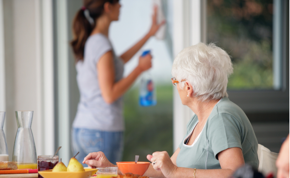 Professional Light Housekeeping Support for Seniors at Home Caregiver providing light housekeeping by cleaning a window while an older woman enjoys breakfast at the table.